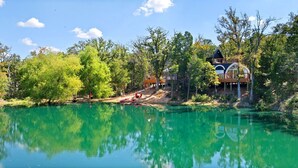 A lakeside property with a house featuring a large dome and a tower, surrounded by trees and a clear lake.