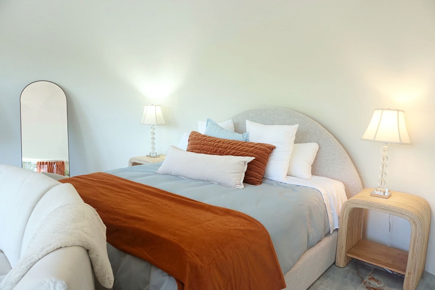 Ein ordentlich bezogenes Bett mit einer braunen Decke, einem grauen Kopfteil und mehreren Kissen. Zwei Nachttische mit Lampen sowie ein großer Spiegel an der Wand sind zu sehen.
