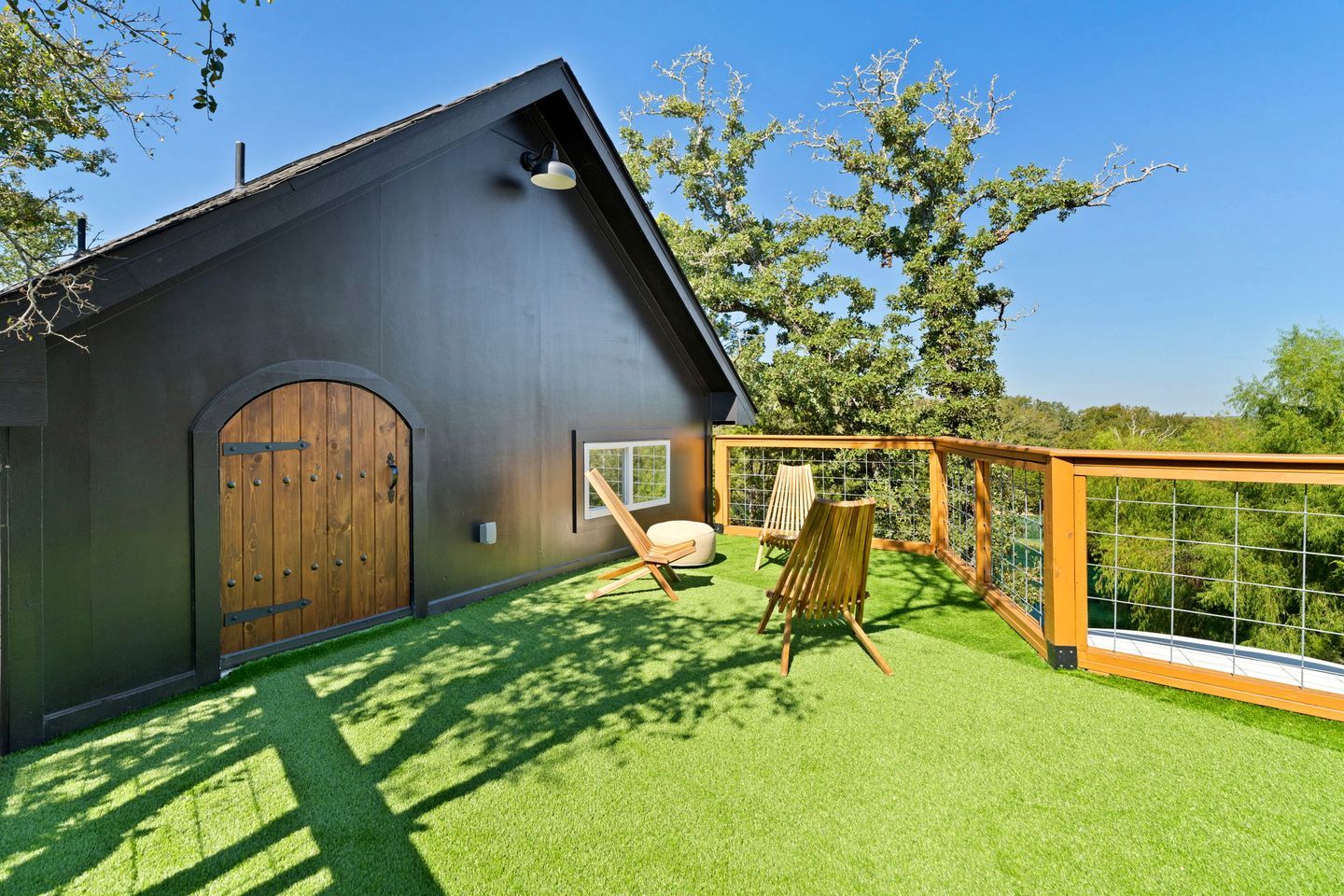 Uma casa moderna com fachada escura, porta de madeira, pequena janela e área externa cercada com duas cadeiras.