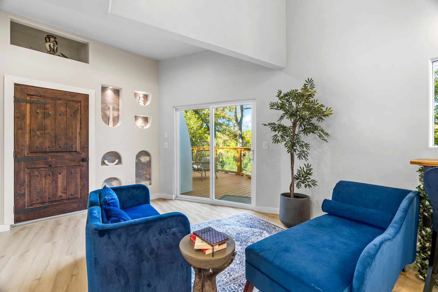 Un salon moderne doté d’une grande porte-fenêtre coulissante donnant sur une terrasse, de canapés bleus, d’une plante en pot et d’un mur décoratif orné de motifs géométriques.