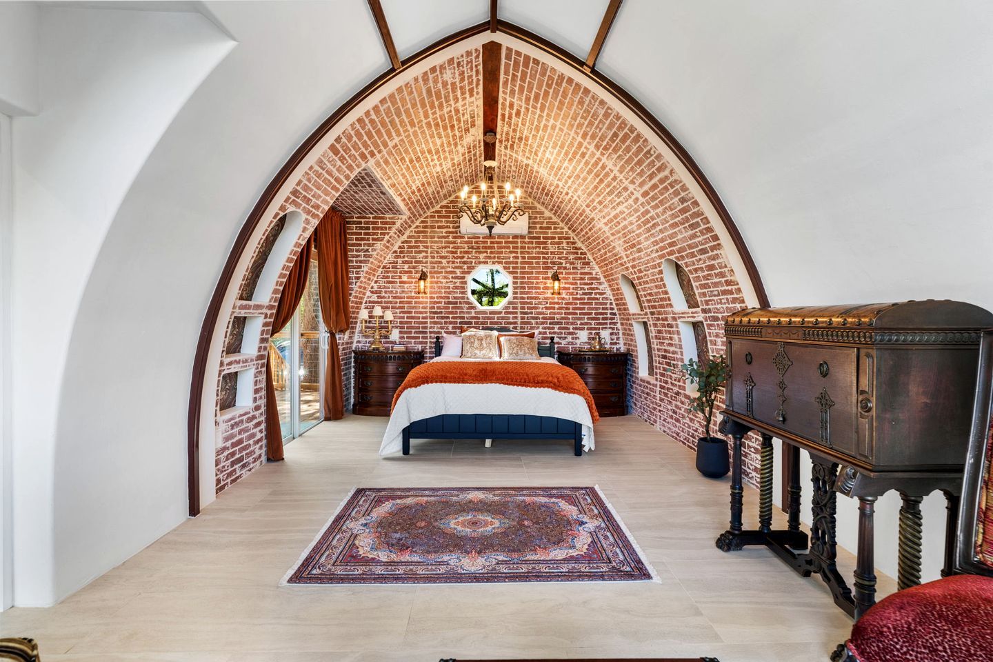 Een slaapkamer met een bakstenen plafond, een groot bed, een kroonluchter en een vintage bijzettafeltje.