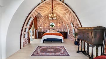 Een slaapkamer met een bakstenen plafond, een groot bed, een kroonluchter en een vintage bijzettafeltje.