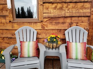 Deux chaises en plastique blanc avec des coussins colorés, une petite table ornée d’un bouquet de fleurs et un mur en bois avec une fenêtre.