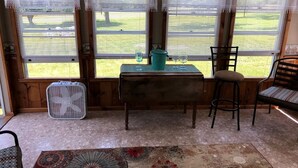 Ein Zimmer mit Tisch, zwei Stühlen, einem Ventilator und Blick auf ein grünes Feld durch Fenster.
