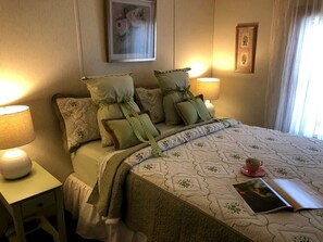 Ein ordentlich bezogenes Bett mit Kissen, eine Nachttischlampe, ein gerahmtes Bild an der Wand und ein Fenster mit Vorhängen.