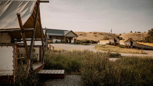 A rustic accommodation with a large tent-like structure, a main building with a sloped roof, and several smaller cabins in a grassy, hilly landscape. - Gorgeous A-Frame Cabin for Two near Yellowstone with Incredible Glampground Amenities in Emigrant, Montana (Emigrant)