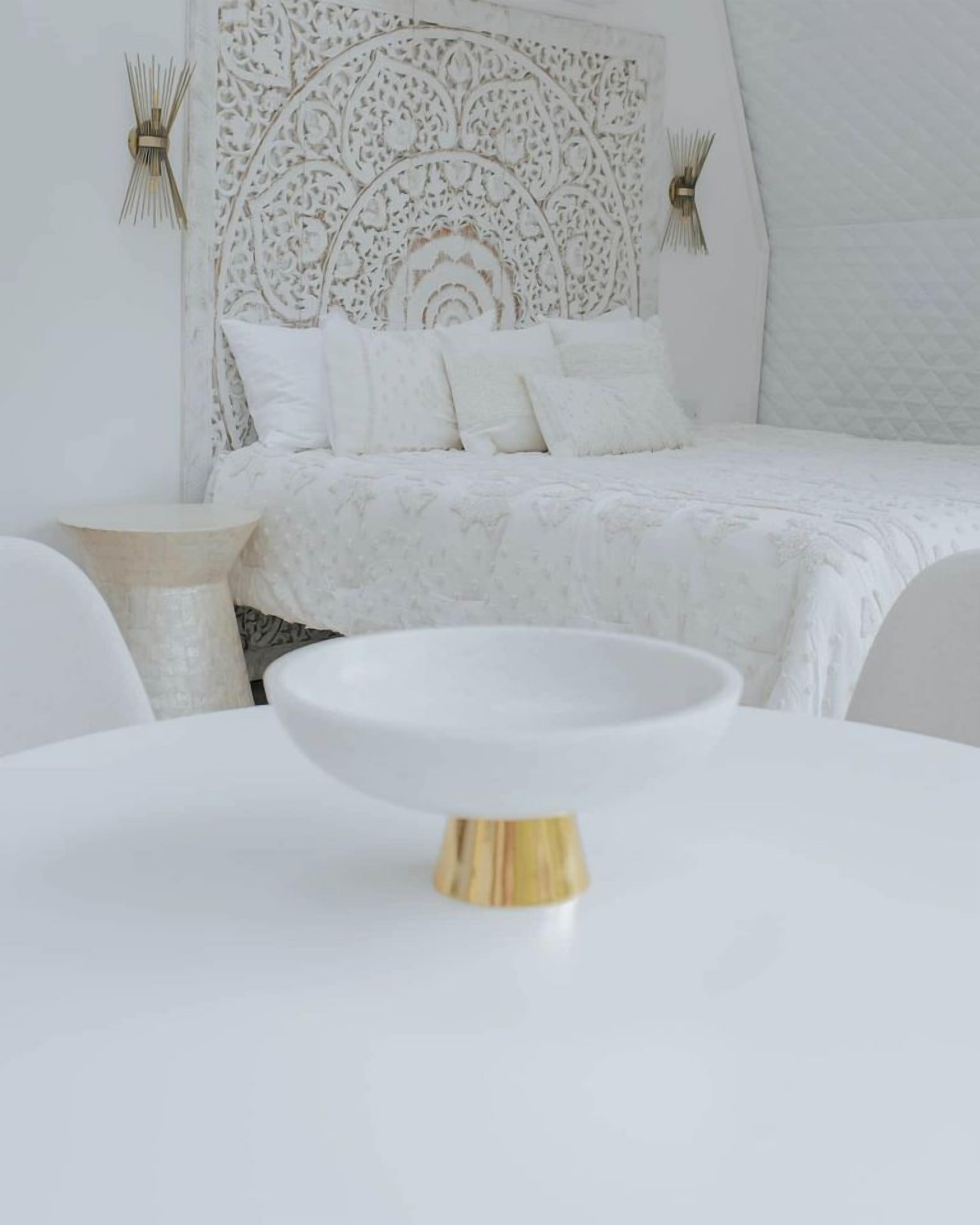 Una stanza bianca con un letto, un tavolino rotondo con una ciotola bianca e una testiera decorativa.