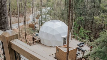 Una estructura de domo geodésico con terraza y zona de estar al aire libre, rodeada de un bosque.
