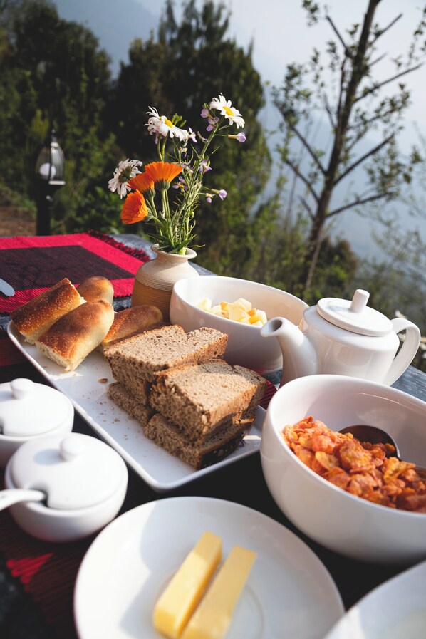 Una mesa con pan, mantequilla, un cebrero y un bol de cereal, ubicada al aire libre con vista a árboles.