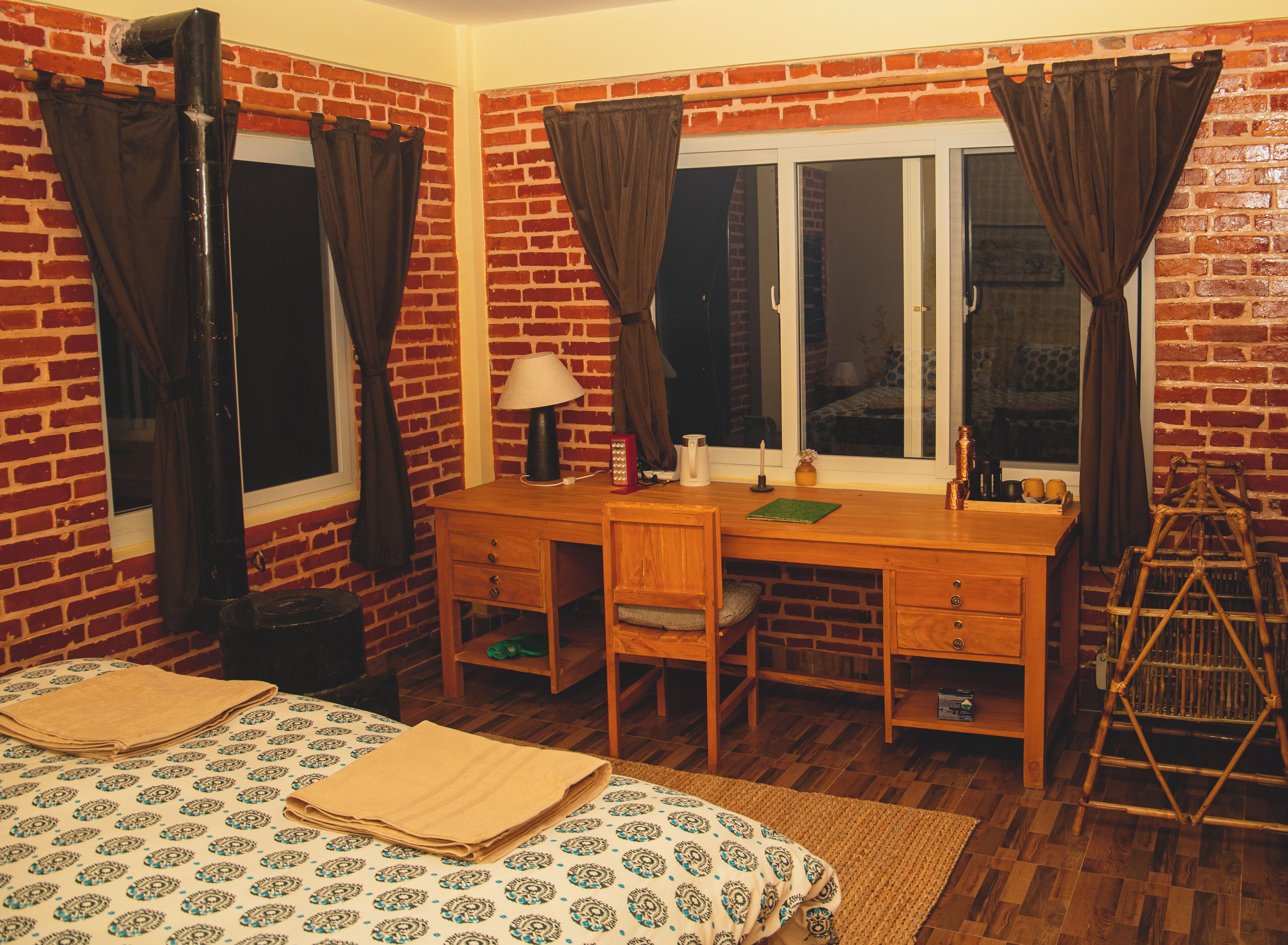 Une pièce avec un mur en briques, un bureau avec une lampe, une chaise, un lit recouvert d’une courtepointe à motifs et une fenêtre avec des rideaux.