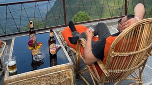 Una persona descansando en una silla de mimbre con vistas a montañas y una barandilla.