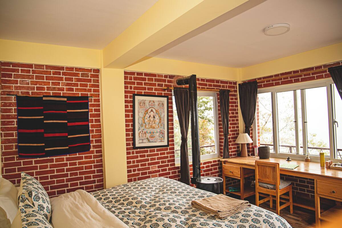 Una habitación con pared de ladrillo, una cama con ropa de cama estampada, un escritorio con silla y una ventana con cortinas.