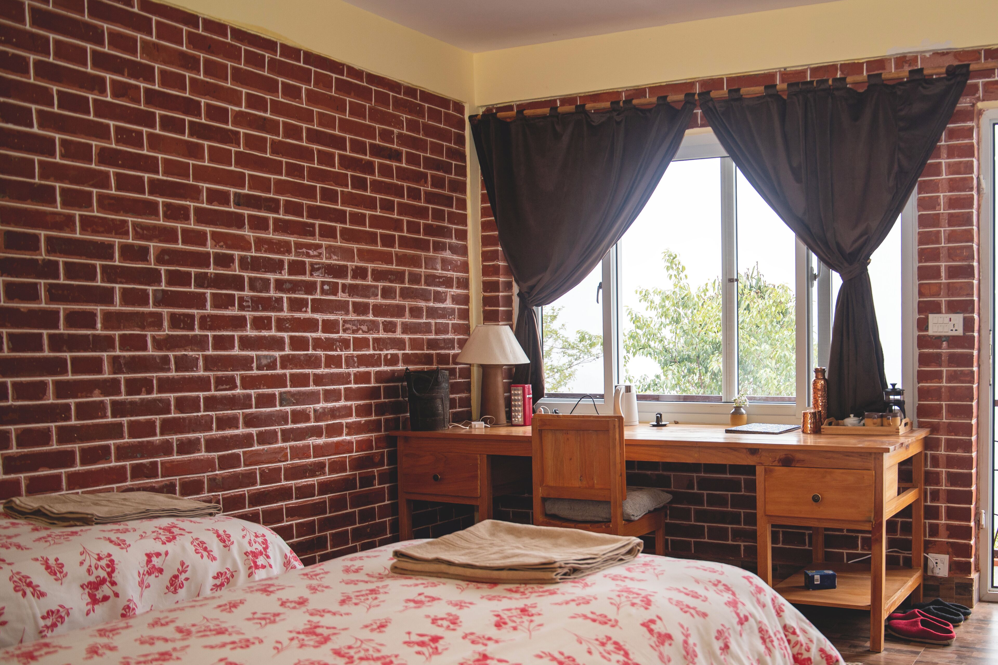 A room with a brick wall, a bed with floral bedding, a desk with a lamp, and a window with curtains.