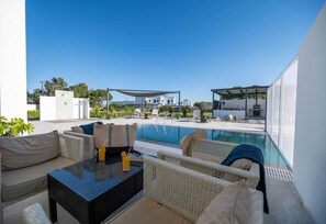 A poolside area with wicker furniture, a glass table, and a view of the pool and buildings in the background. - Vigla Kiotari - A, Villa (Greece)