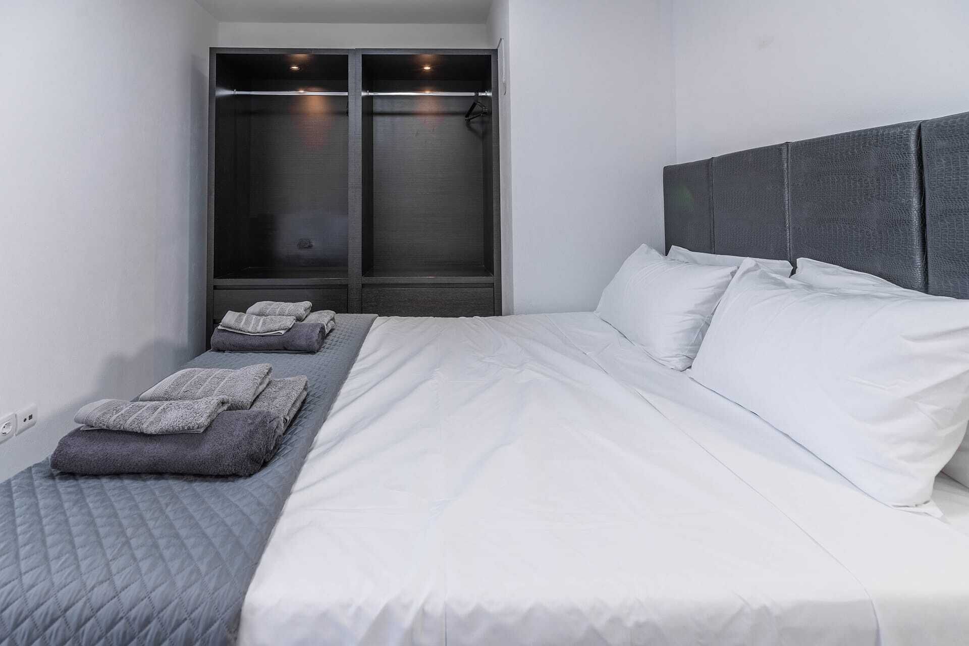 Un dormitorio con cama, almohadas, cabecera, mesita de noche, estante en la pared y armario.