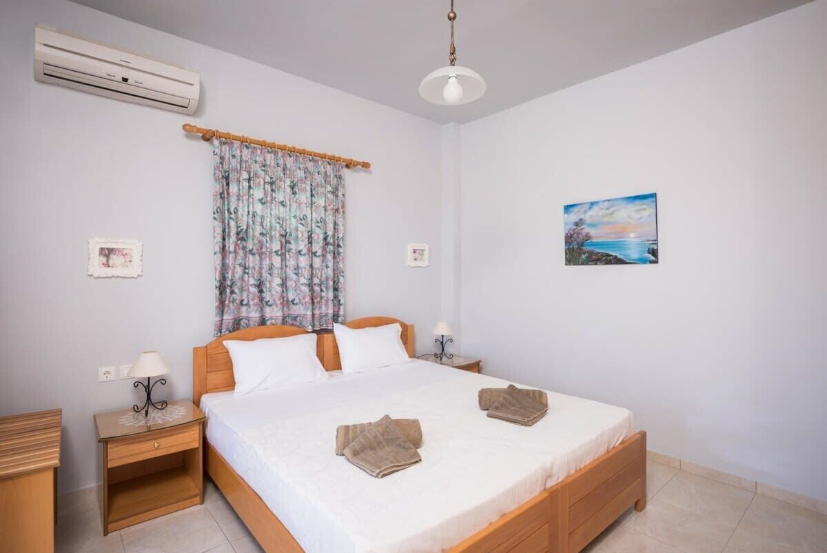 A neatly arranged bedroom with a bed, bedside tables, an air conditioner, a curtain, and a wall-mounted picture.