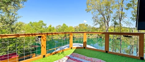 A balcony with a view of a lake, surrounded by trees, and a patterned rug on the floor.