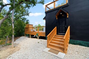 Un bâtiment noir avec un escalier en bois menant à une porte noire, le tout entouré de gravier et d’arbres.