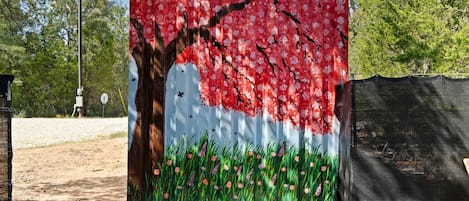Un mural grande y colorido en una valla que muestra un árbol con flores rosadas y césped verde.