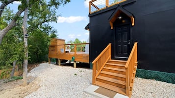 A black building with a wooden staircase leading to a black door, surrounded by gravel and trees.