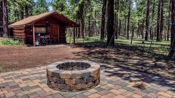 Uma cabana rústica com uma lareira de fogo em primeiro plano, cercada por uma área arborizada.