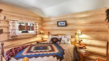 Quarto de cabana de madeira com cama, duas mesas de cabeceira, janela com cortinas e um quadro na parede.