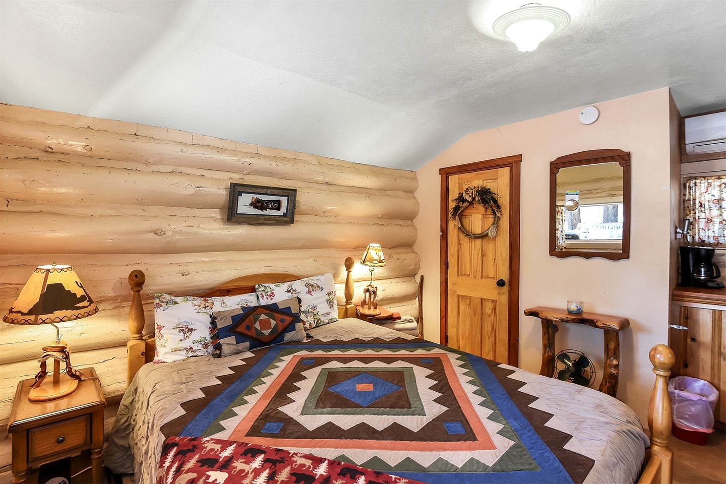 Quarto aconchegante com parede de madeira, cama com colcha estampada, abajures, mesa de cabeceira de madeira, porta com grinalda e espelho na parede.