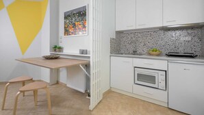 Ett kompakt kök med vita skåp, en bänkskiva med ett mönstrat stänkskydd, en inbyggd ugn och en liten matplats med två barstolar.
