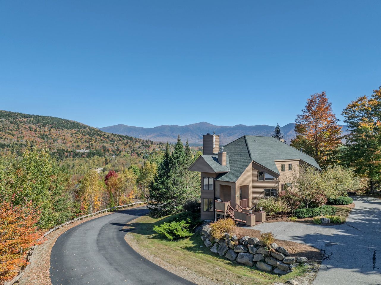 Une maison avec une allée sinueuse, entourée d’un feuillage automnal et avec des montagnes en arrière-plan.