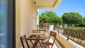 A balcony with a wooden table and chairs, a small table with a basket and a pitcher, and a view of a garden with trees and a fence. - Rodini Anassa | Apollo, Apartment (Rodos)