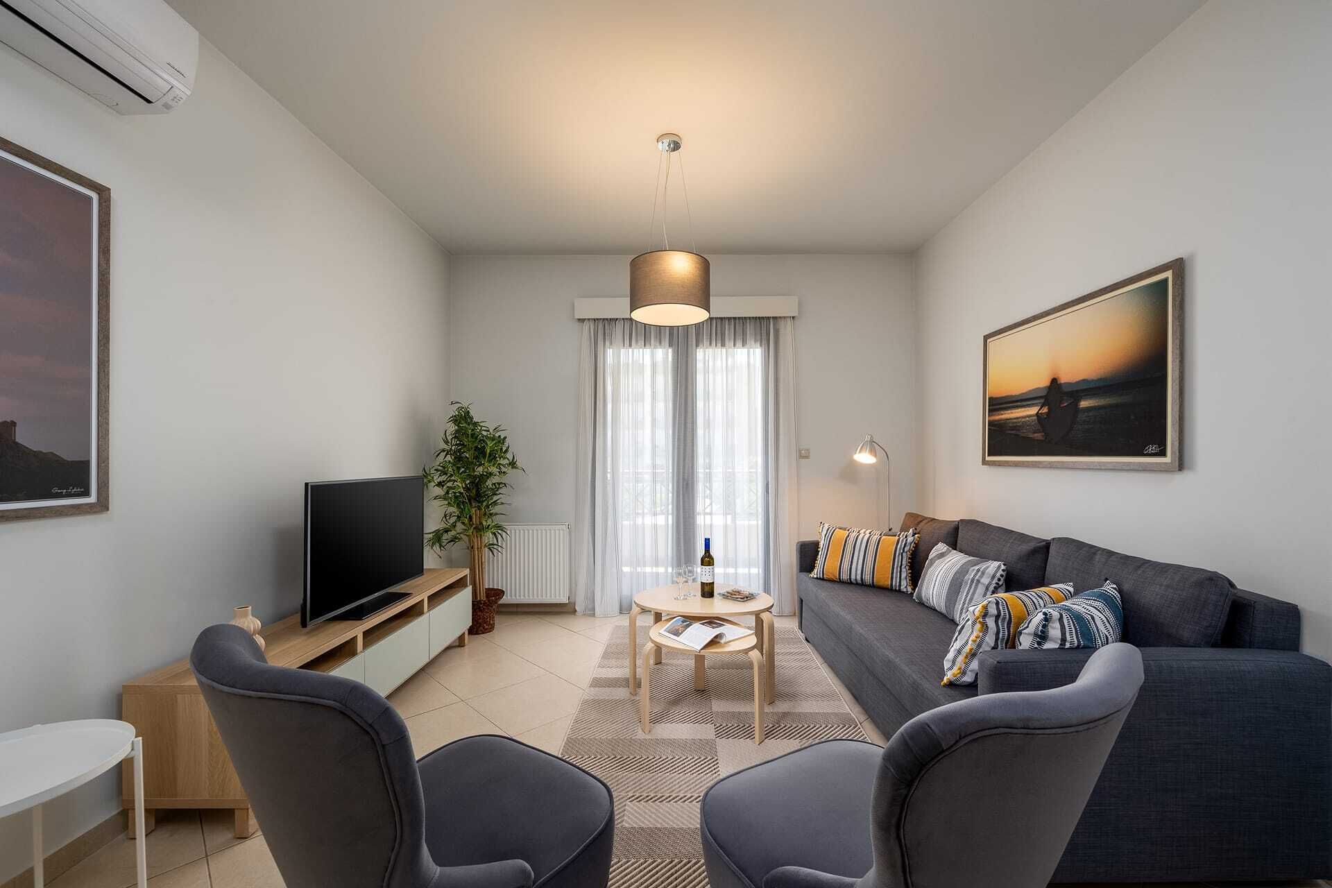 Ein modernes Wohnzimmer mit einer Couch, zwei Sesseln, einem Fernseher, einem Couchtisch und einem großen Fenster mit Vorhängen.