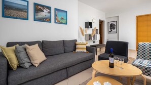 Een woonkamer met een grijze bank, een ronde houten salontafel en een zwarte fauteuil. Er hangen drie ingelijste foto's aan de muur, een klok en een deur.