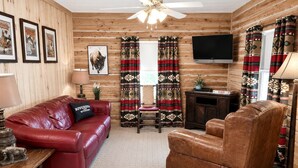 A log cabin interior with a red leather sofa, a brown leather armchair, a wooden coffee table, a television mounted on the wall, and patterned curtains.