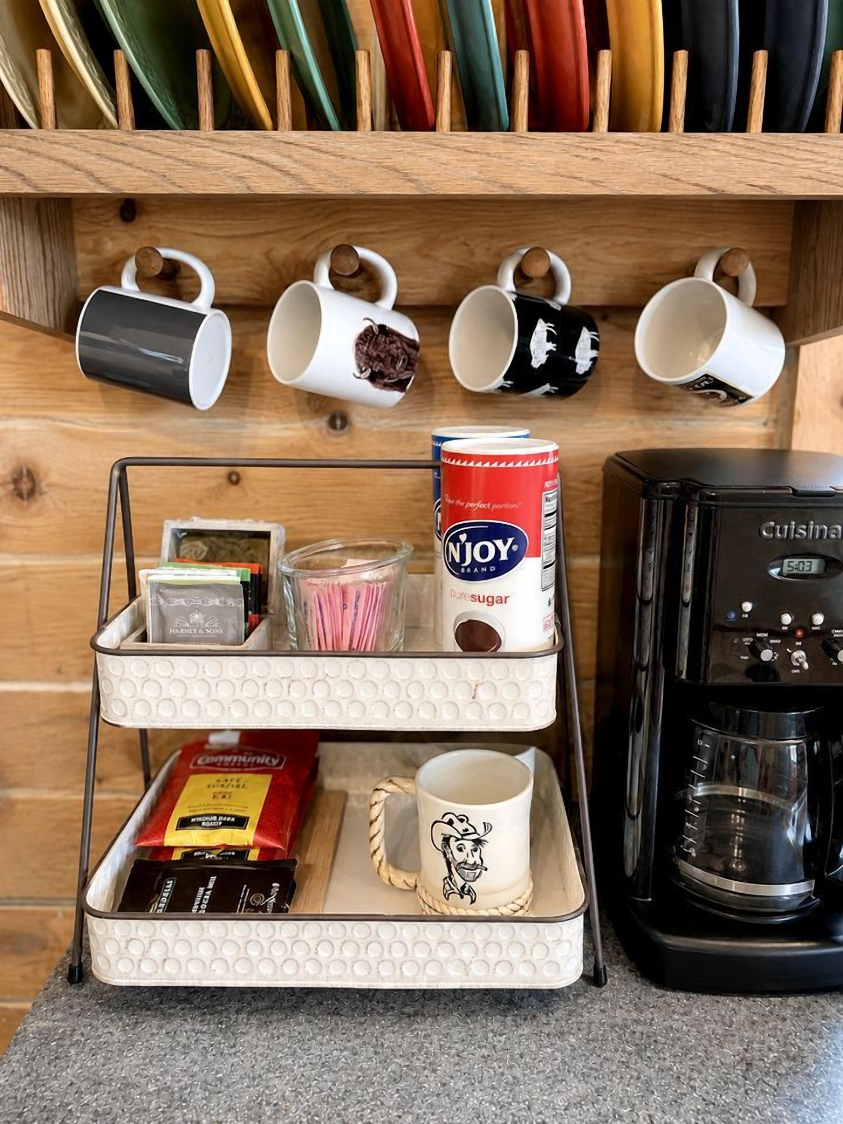 A kitchen counter with a coffee maker, a mug rack, and a tray with coffee pods and a can of sugar.