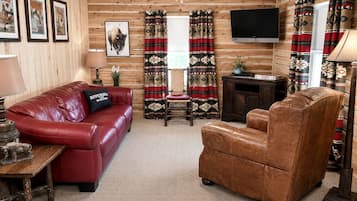 Een interieur van een blokhut met een rode leren bank, een bruine lederen fauteuil, een houten bijzettafeltje, een aan de muur bevestigde televisie en raambekleding met een patroon.