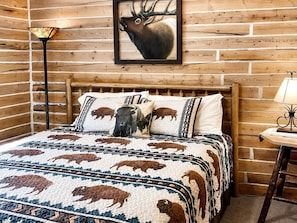 A log cabin-style room with a wooden bed, buffalo-themed bedding, a framed elk painting, and a lamp. - Tranquil Two Bedroom Cabin in Riverside, Wyoming (Riverside)