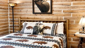 A log cabin-style room with a wooden bed, buffalo-themed bedding, a framed elk painting, and a lamp.