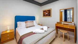 A hotel room with a bed, a wooden nightstand, a desk, a chair, a mirror, and a framed picture on the wall. - Rodini Anassa | Dimitra, Apartment (Rodos)