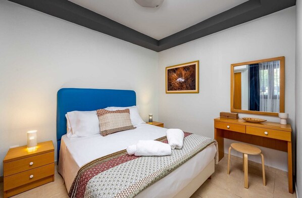 Habitación de hotel con cama, mesita de noche de madera, escritorio, silla, espejo y un cuadro en la pared.