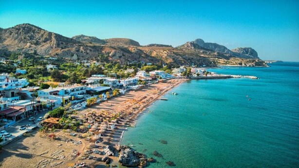 A coastal town with buildings along the beach, clear blue waters, and rocky hills in the background. - Summer Paradise Stegna (RODOS)