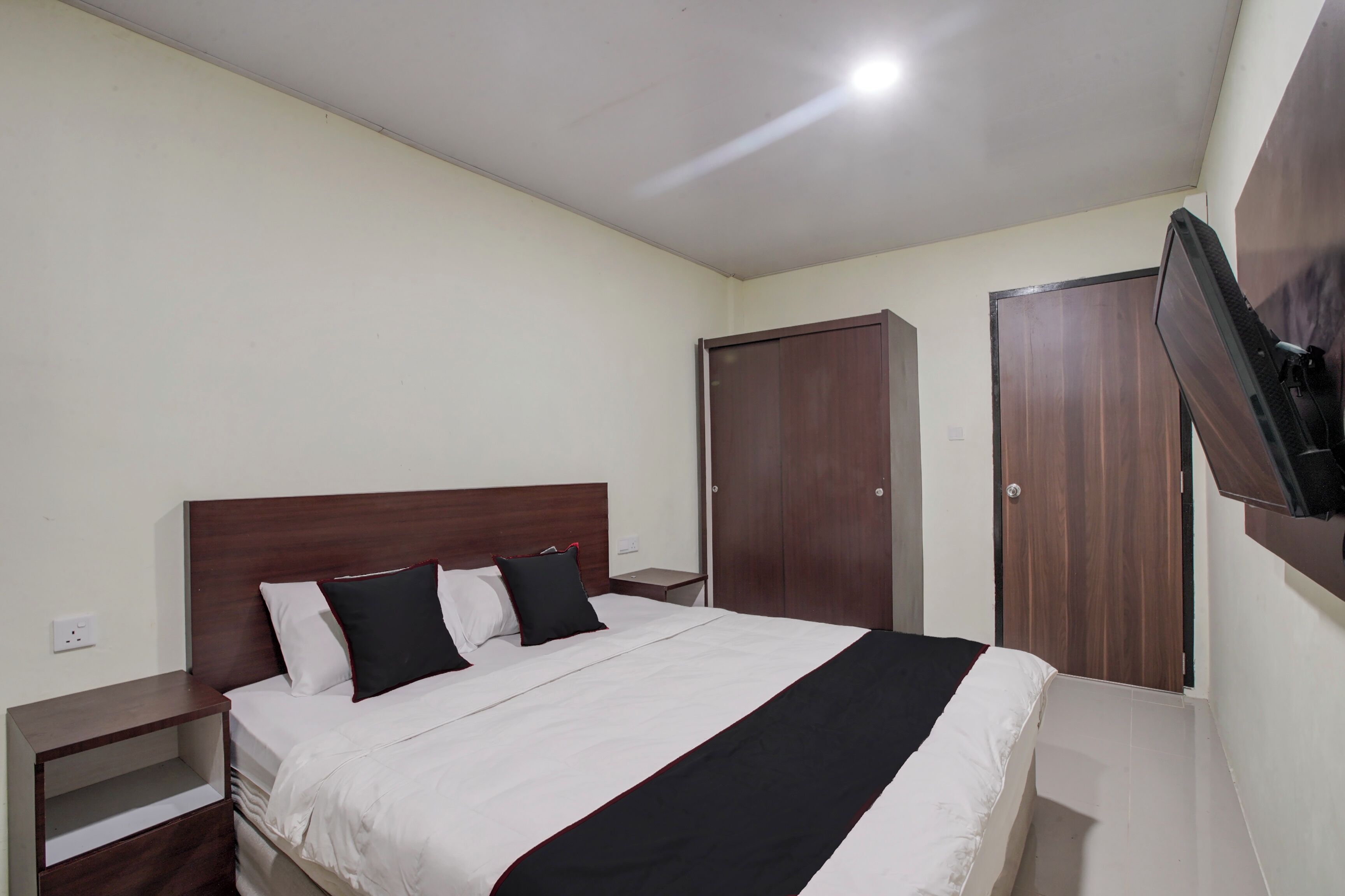 A hotel room with a bed, a nightstand, a TV mounted on the wall, and two wooden doors.
