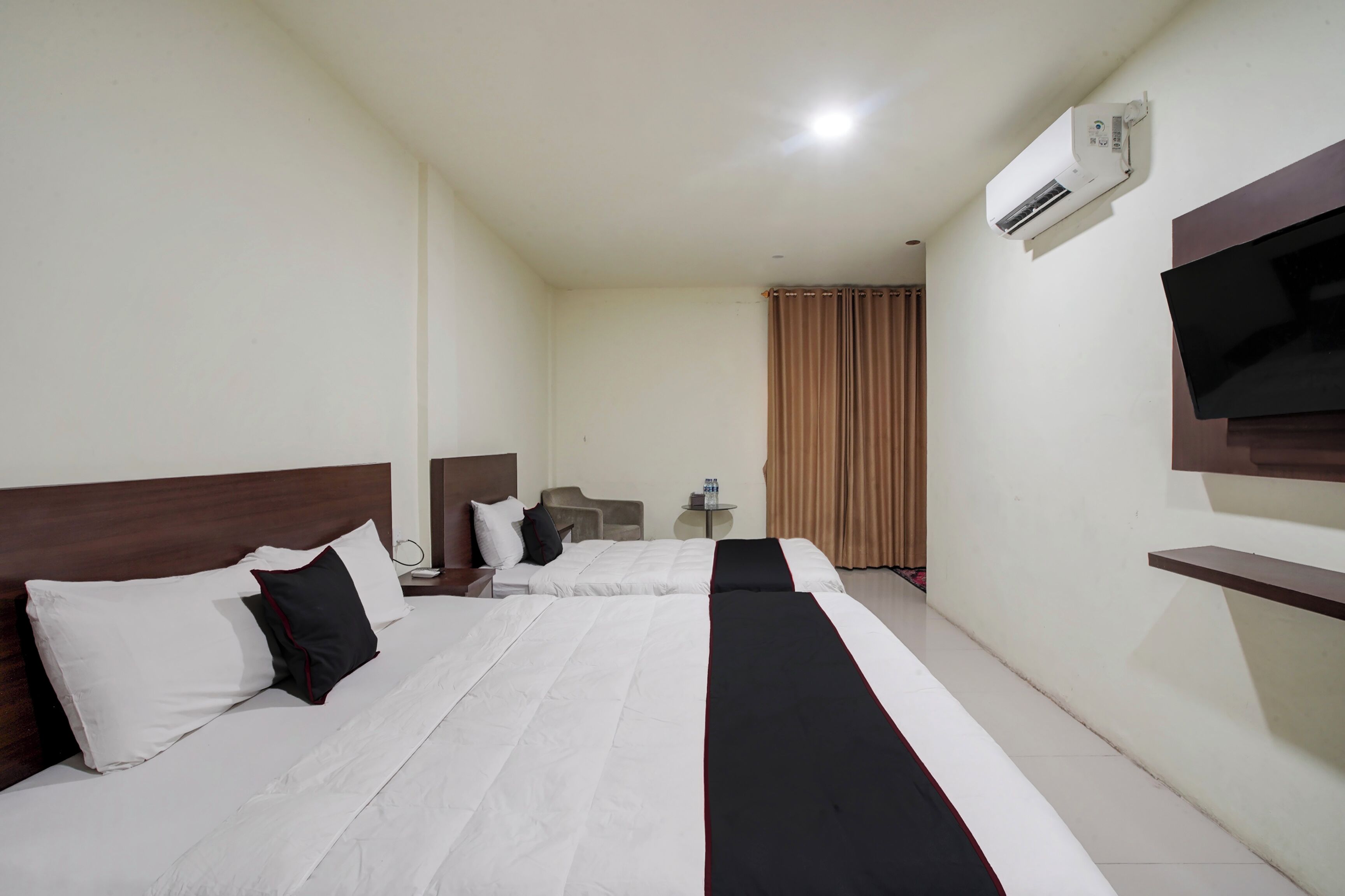 Une chambre d’hôtel avec deux lits, une télévision, un appareil de climatisation, une petite table avec une bouteille et un rideau.