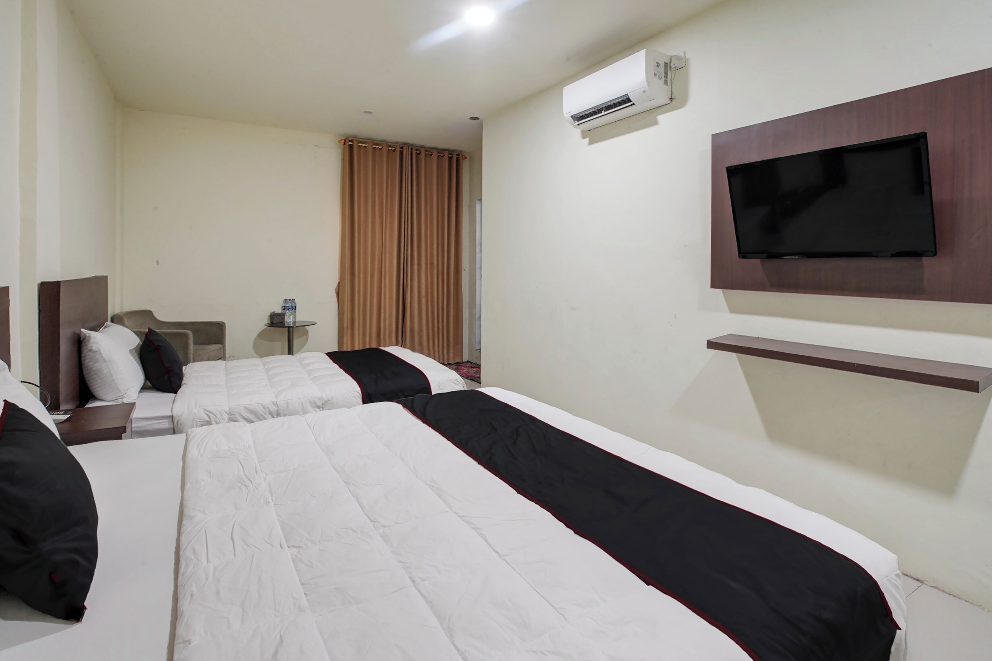 A hotel room with two beds, a TV mounted on the wall, an air conditioning unit, and a small table with a bottle.