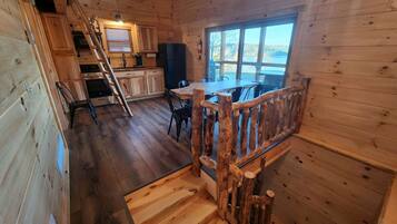 A wooden cabin interior with a staircase, kitchen, dining area, and a large window offering a view.