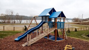 Un parque infantil con tobogán y estructuras para escalar, rodeado por una cerca y una masa de agua al fondo.