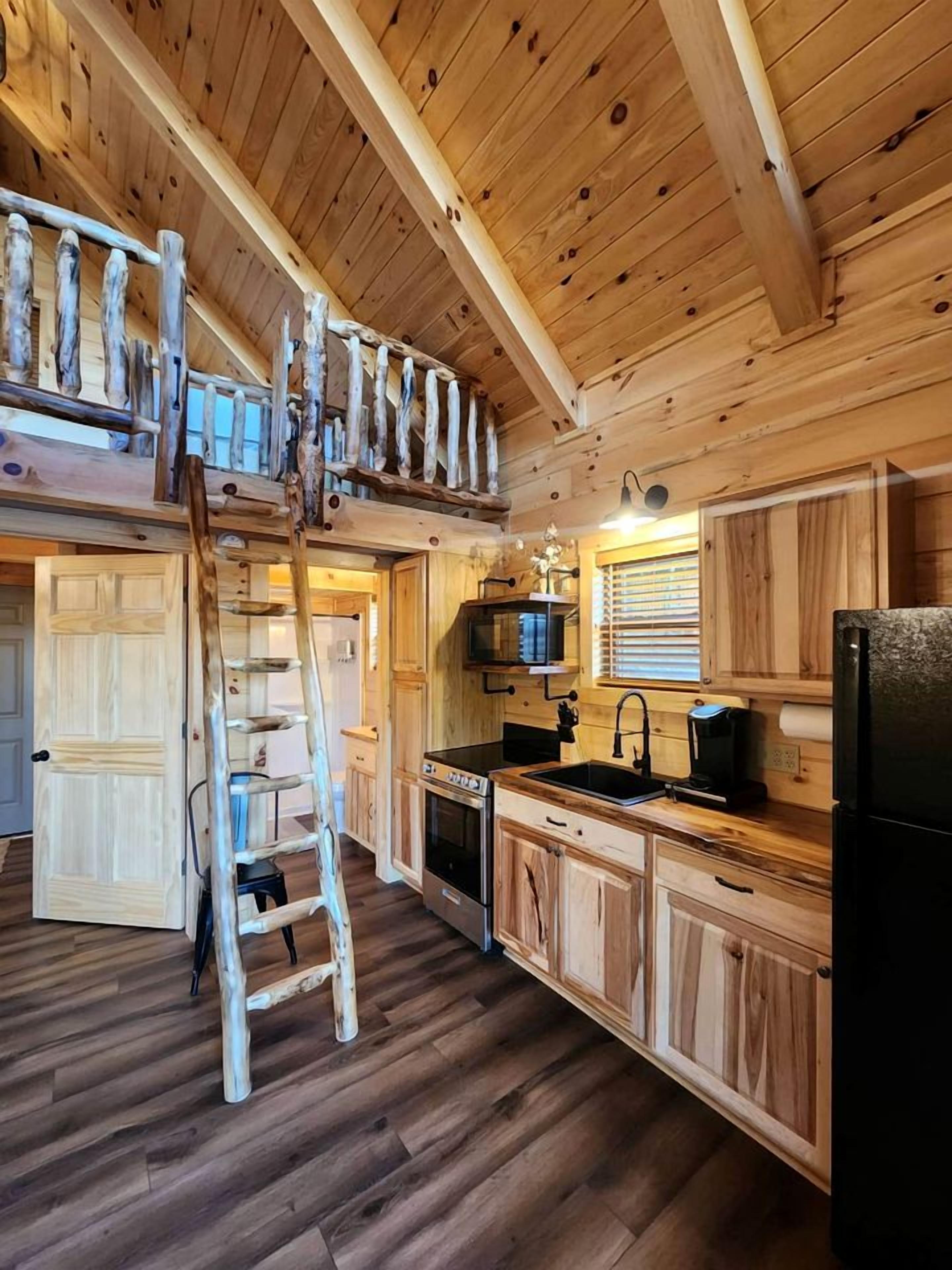 A wooden cabin interior with a loft bedroom, a kitchen with wooden cabinets, a stove, and a refrigerator.