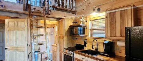 A wooden cabin interior with a loft bedroom, a kitchen with wooden cabinets, a stove, and a refrigerator.