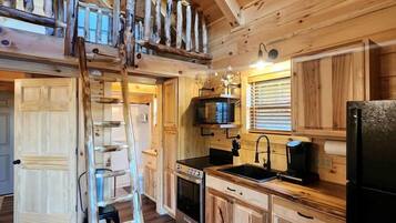 A wooden cabin interior with a loft bedroom, a kitchen with wooden cabinets, a stove, and a refrigerator.