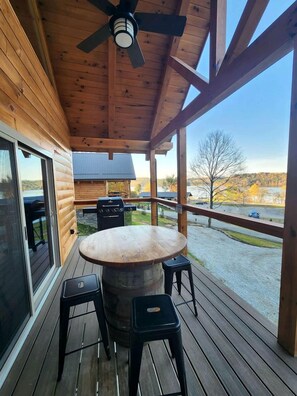 A wooden deck with a round table and two stools, a ceiling fan, and a view of a lake and trees. - Group Friendly Cabin with Spa on the Deck near Tappan Lake in Ohio (Deersville)
