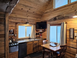 A compact kitchen with wooden walls, a dining area, and a television mounted above the sink.